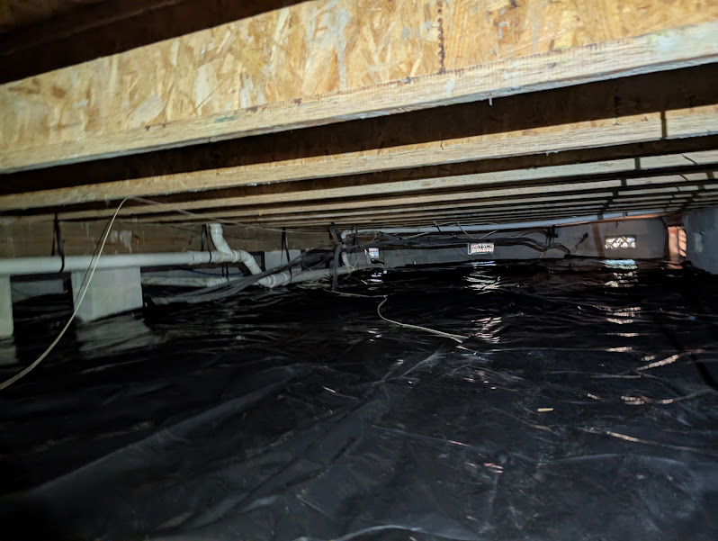Black vapor barrier laid tight across a low crawlspace floor beneath plumbing and wiring